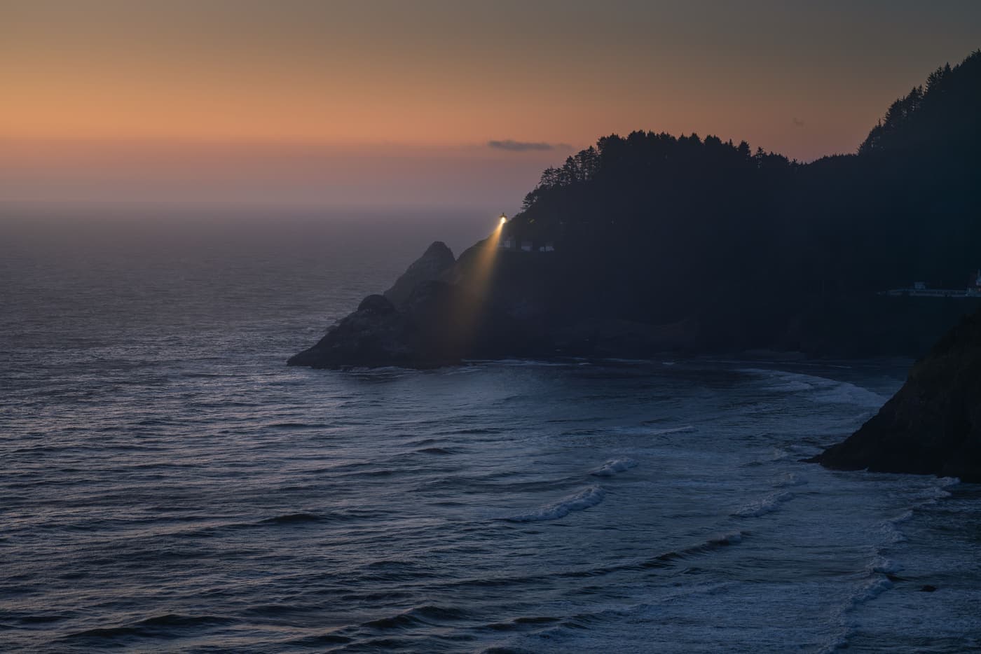 Lighthouse beam sweeping across the sea from a cliff at dusk