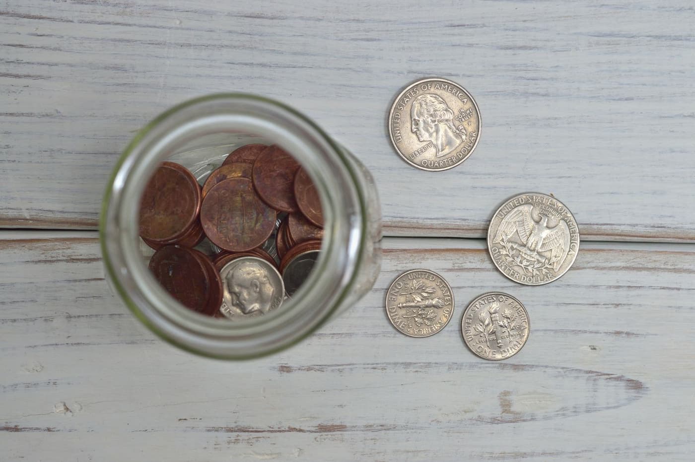 A glass jar of coins on a wooden table in daylight.
